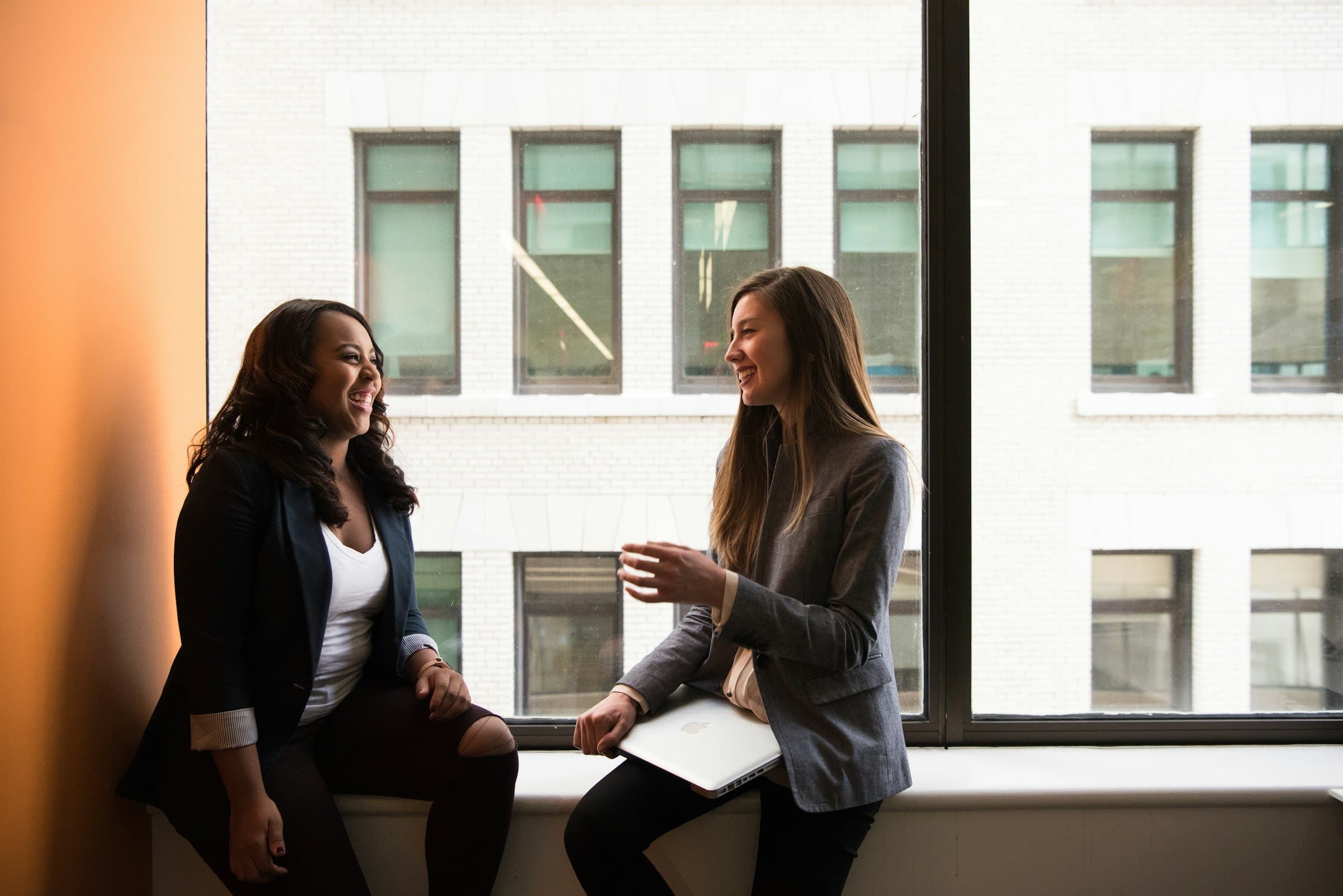 Two women laugh together by an office window.